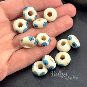 以下が含まれることがあります： 青い邪悪な目のデザインが施された白いセラミックビーズのコレクション。ビーズの中央には、糸を通すための穴があります。