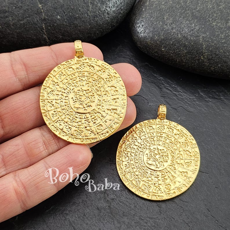May include: Two gold-toned round pendants with intricate circular designs and raised lettering. The pendants are approximately 2 inches in diameter.