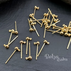 Könnte beinhalten: Nahaufnahme eines Stapels goldfarbener Ohrring-Stifte mit kleinen runden Kugeln an den Enden. Die Stifte sind auf einer schwarzen Oberfläche verstreut.