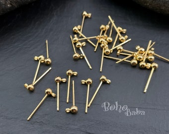 May include: A close-up of a pile of gold-coloured earring posts with small round balls on the ends. The posts are scattered on a black surface.
