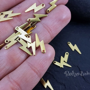 May include: A close-up of a hand holding a pile of gold-coloured lightning bolt charms. The charms are made of metal and have a shiny finish. The charms are perfect for adding a touch of whimsy to jewellery or crafts.