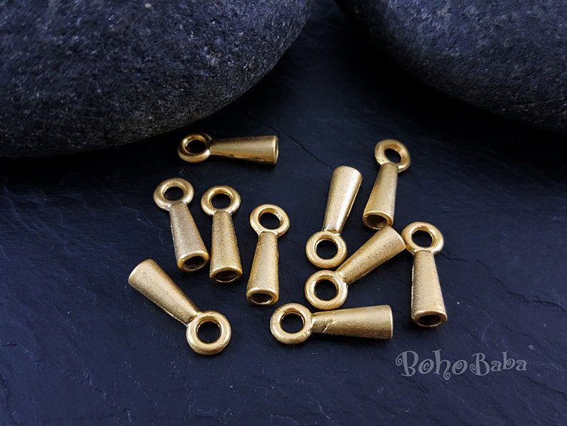 May include: A group of 10 gold-toned metal cord end caps with a loop at the top. The cord end caps are approximately 2.5 cm long and have a tapered shape.