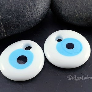 May include: Two white ceramic evil eye charms with blue and black details. The charms are oval shaped and have a hole at the top for hanging.