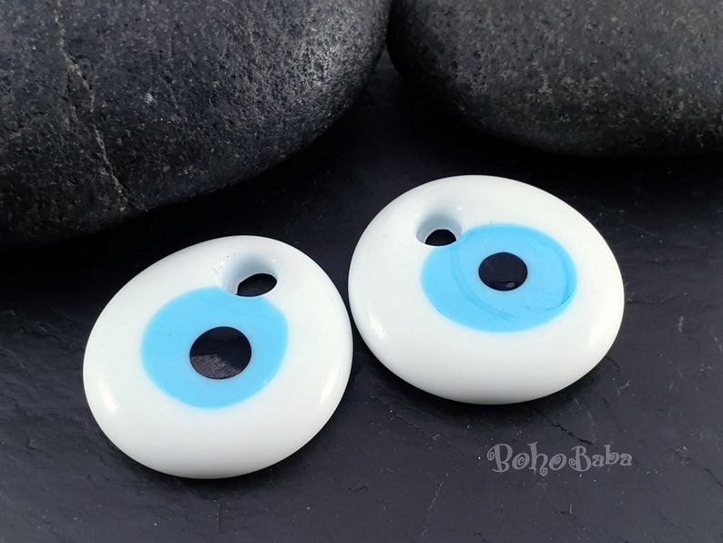 May include: Two white ceramic evil eye charms with blue and black details. The charms are oval shaped and have a hole at the top for hanging.
