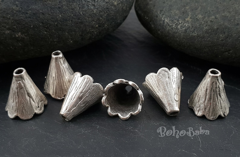 May include: Six silver metal cone-shaped beads with scalloped edges. The beads are arranged in a semi-circle on a dark gray surface.