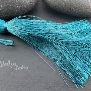 May include: Teal blue tassel with a long, flowing fringe. The tassel is made of soft, silky threads and has a small, round knot at the top.