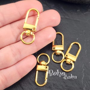 May include: Gold-colored swivel clasps, perfect for jewelry making or crafting. The clasps have a rectangular base with a rounded top and a circular ring for attaching to other components. The image shows several clasps, some held in a hand.