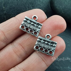 May include: Two silver-toned metal charms with a geometric design and a loop at the top. The charms have a repeating pattern of circles, lines, and diamonds.