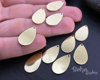 May include: Ten gold-colored metal teardrop-shaped charms with a hole at the top. The charms are arranged on a hand and a dark surface.