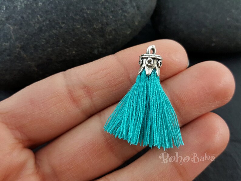May include: Teal tassel with a silver metal top. The tassel is made of soft, thin strands of yarn and has a decorative, ornate metal top.