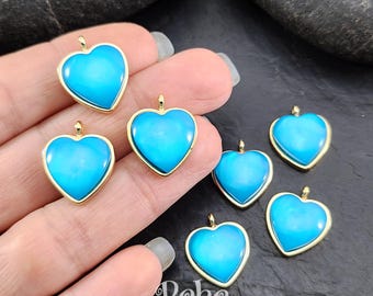Puede incluir: Siete dijes en forma de corazón de tono dorado con un centro azul turquesa. Los dijes están esparcidos sobre una superficie oscura.