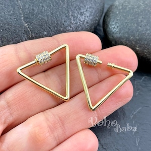 May include: A pair of gold triangle earrings with a clear crystal accent on the hinge. The earrings are shown on a hand.
