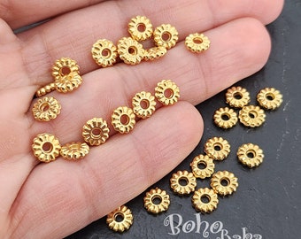 May include: A close-up of a hand holding a large quantity of gold-colored metal beads. The beads are round with a scalloped edge and have a hole in the center. The beads are arranged in a random pattern on the hand. The background is a dark gray surface with the text "boho baba" printed in white.