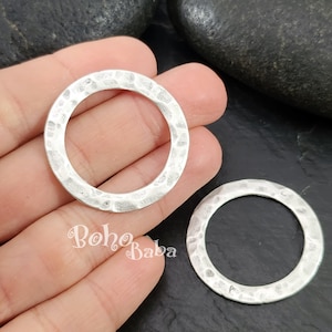 Pode incluir: Dois anéis de metal martelado prateados. Os anéis são redondos e têm um acabamento liso e polido. Eles medem aproximadamente 5 cm de diâmetro.