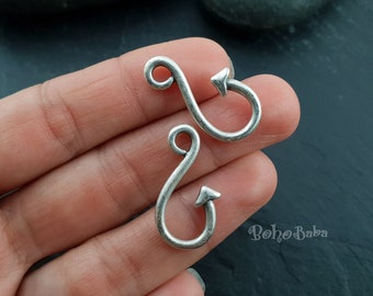 May include: Two silver-toned metal clasps with a curved design and arrow-shaped ends. The clasps have a vintage, antique appearance. The background is a dark, textured surface. The Boho Baba logo is visible.