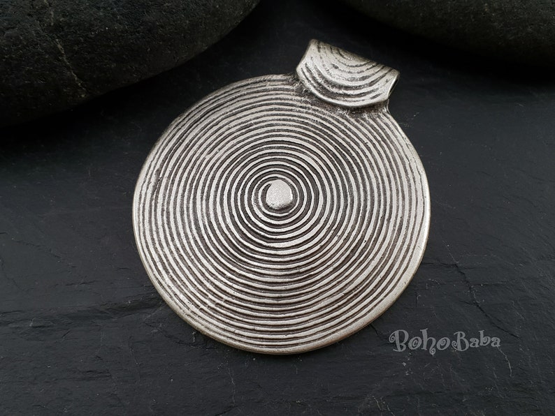 May include: Silver pendant with a circular design featuring concentric circles. The pendant has a loop at the top for attaching to a chain or cord.