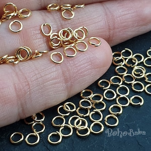 May include: A close-up of a large quantity of gold jump rings. The rings are small and round, and they are scattered on a surface. The rings are used for jewelry making.