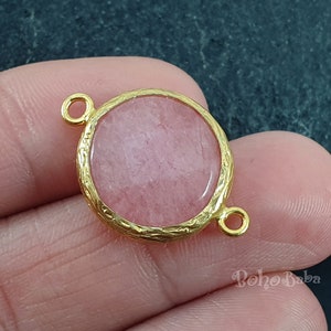 Può includere: Un ciondolo in metallo color oro con un centro in pietra rosa rotonda. Il ciondolo ha un bordo strutturato e piccoli anelli su ogni lato per il fissaggio a una catena o ad un altro componente di gioielleria. Il ciondolo è tenuto in mano.