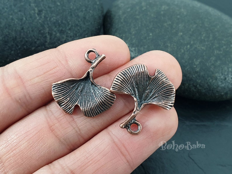 May include: Two copper-colored ginkgo leaf charms. Each charm features a detailed leaf design with a small loop for attaching to jewelry. The charms have a vintage, antique appearance.