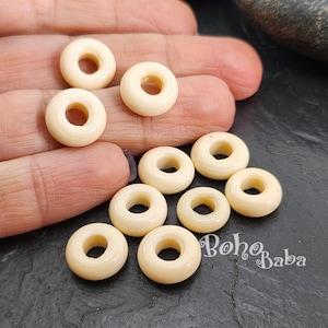 以下が含まれることがあります： 中心に穴の開いたドーナツ型のオフホワイトのプラスチックビーズが10個。ビーズは暗い灰色の表面に半円形に配置されています。