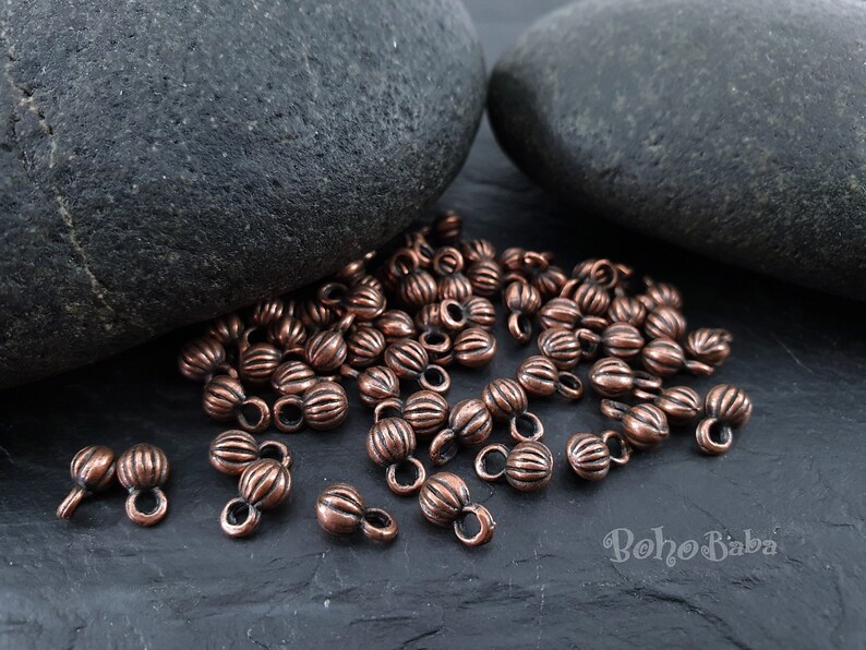 May include: A close-up of a pile of copper-colored metal beads with a ribbed texture. Each bead has a small loop on either end. The beads are scattered on a dark gray surface.