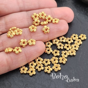 May include: A close-up of a pile of gold-colored metal flower-shaped beads. The beads have a textured surface and a hole in the center. The beads are scattered on a dark surface.
