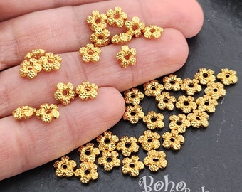 May include: A close-up of a pile of gold-colored metal flower-shaped beads. The beads have a textured surface and a hole in the center. The beads are scattered on a dark surface.