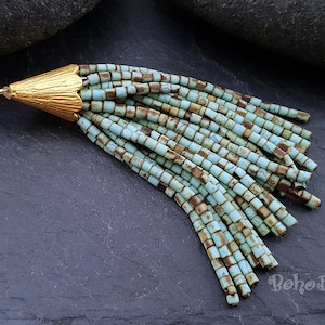 May include: A turquoise and brown beaded tassel with a gold metal top. The tassel is hanging from a dark grey surface.