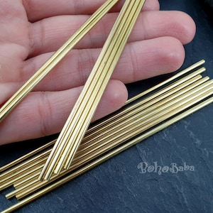 Puede incluir: Un paquete de 12 varillas de latón dorado, de aproximadamente 15 cm de largo, se muestran sobre una superficie gris oscuro. Las varillas están dispuestas en forma de abanico, con las puntas de las varillas apuntando hacia el lado derecho de la imagen. Las varillas son lisas y brillantes, y tienen un borde ligeramente redondeado.