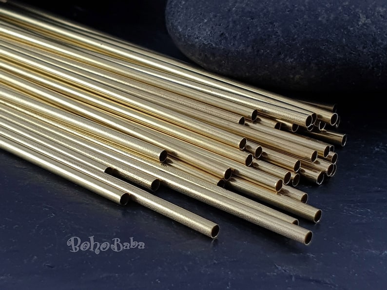 May include: A pile of gold-colored metal tubes, likely for jewelry making, on a dark blue surface. The tubes are arranged in a staggered pattern, with some of the tubes extending beyond the edge of the pile. The tubes are all the same size and shape, and they have a smooth, polished finish.