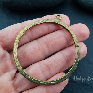 Puede incluir: Un colgante circular de metal color latón antiguo con una superficie texturizada. El colgante tiene un pequeño lazo en la parte superior para colgar y se sostiene en una mano. El diseño general es rústico y bohemio.