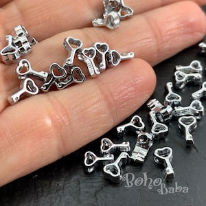 Puede incluir: Primer plano de un montón de dijes de llave plateados con mirillas en forma de corazón. Los dijes son pequeños y delicados, y serían perfectos para agregar un toque de fantasía a las joyas o las manualidades.