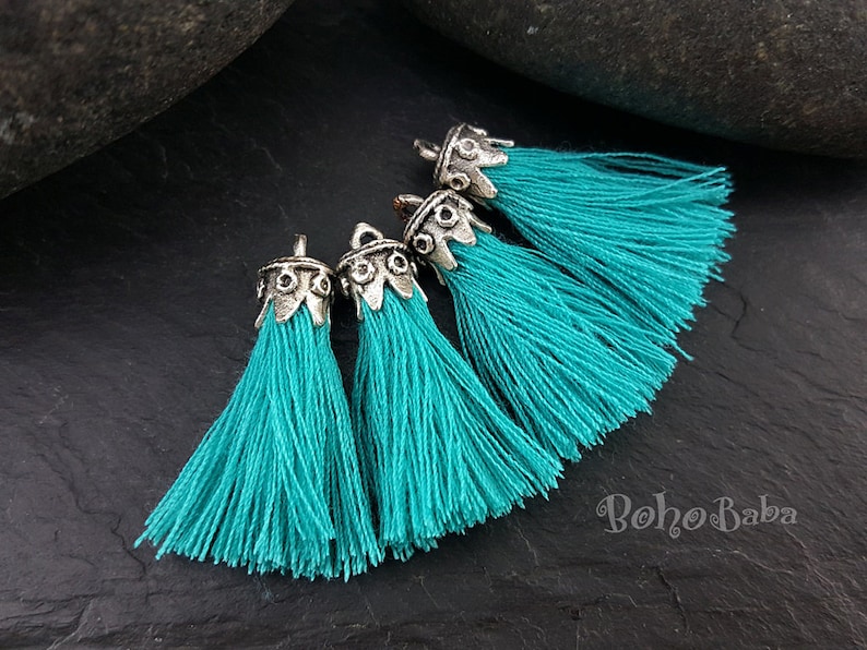 May include: Four turquoise blue tassels with silver metal tops. The tassels are arranged in a semi-circle on a dark gray surface.