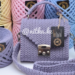 May include: A lavender crocheted handbag with a silver clasp and a long strap. The bag is surrounded by balls of yarn in various colors.