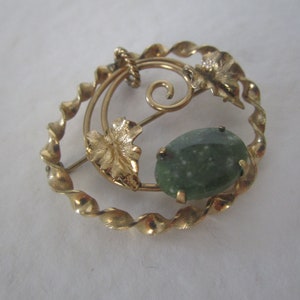 May include: A gold-toned brooch with a green stone set in a twisted metal frame. The frame is decorated with gold leaves.