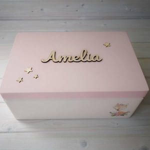 Pode incluir: Uma caixa de madeira rosa com uma base branca. A tampa tem uma borda rosa claro e uma placa de nome de madeira que diz "Amelia". Há pequenas estrelas douradas na tampa e uma pequena ilustração de uma raposa na frente da caixa.