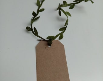 Könnte beinhalten: Ein braunes Papier-Geschenk-Etikett mit einer grünen Blättergirlande, die um das obere Loch gebunden ist.