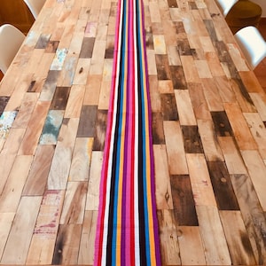 May include: A colorful striped table runner with black, white, pink, yellow, blue, and orange stripes. The runner is laid out on a wooden table with a rustic, reclaimed wood look.