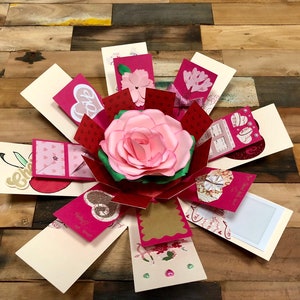 Op de afbeelding: Een roze en rode explosiebox met een roze papieren roos in het midden. De doos is versierd met harten, glitter en andere versieringen. De doos is open en de zijkanten zijn uitgeklapt om de verschillende compartimenten te laten zien.