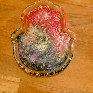 Può includere: Un vassoio di vetro trasparente a forma di mano di hamsa con un design in resina colorata e scaglie dorate. Il vassoio ha un design a mandala al centro.