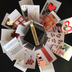 May include: A white and red explosion box with a black ring box in the centre. The box is decorated with hearts, stars, and the words "Love you" and "Will you marry me?"