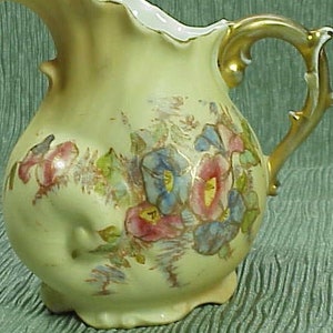 May include: A vintage, pale yellow ceramic pitcher with a ruffled rim and a gold-colored handle. The pitcher is adorned with hand-painted floral designs in shades of pink, blue, and green. The pitcher has a bulbous body and a small, decorative foot.