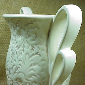 May include: White ceramic vase with an intricate floral design and two handles. The vase has a smooth, glossy finish.