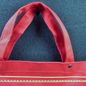 Puede incluir: Bolsa de lona roja con dos asas y ribete dorado. La bolsa tiene tres remaches met&aacute;licos.