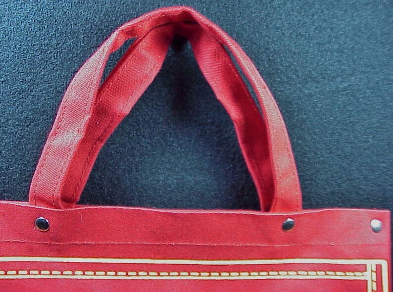 Puede incluir: Bolsa de lona roja con dos asas y ribete dorado. La bolsa tiene tres remaches met&aacute;licos.