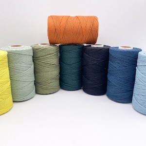 May include: A selection of macrame cord spools in a range of colours. The spools include shades of green, blue, and orange. The cords are tightly wound, ready for crafting. The image is set against a white background.