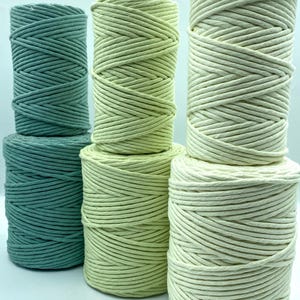 May include: A selection of macrame cord spools in teal, light green, and cream. The cords are tightly wound, creating a textured look. The spools are arranged in a visually appealing composition, ideal for various crafting projects.