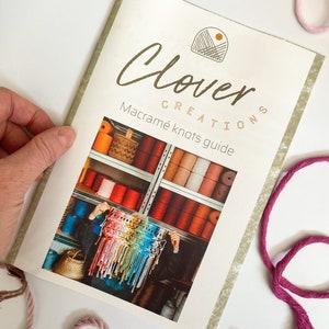 May include: A book titled "Clover Creations Macrame Knots Guide" with a green and white striped cover. The cover features a hand-drawn logo of a sun and a house with a woven pattern. The book is open to a page with a photo of a person holding a colorful macrame wall hanging in front of a shelf filled with colorful yarn.