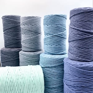 May include: A selection of cotton rope spools in shades of blue and grey. The colours vary from light teal to deep indigo. The rope is tightly wound, creating a textured surface. The spools are arranged in a visually appealing display.
