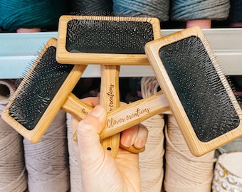 May include: Three wooden brushes with black bristles and the text "Clover creations" engraved on the handle. The brushes are held in a hand with a tattoo.